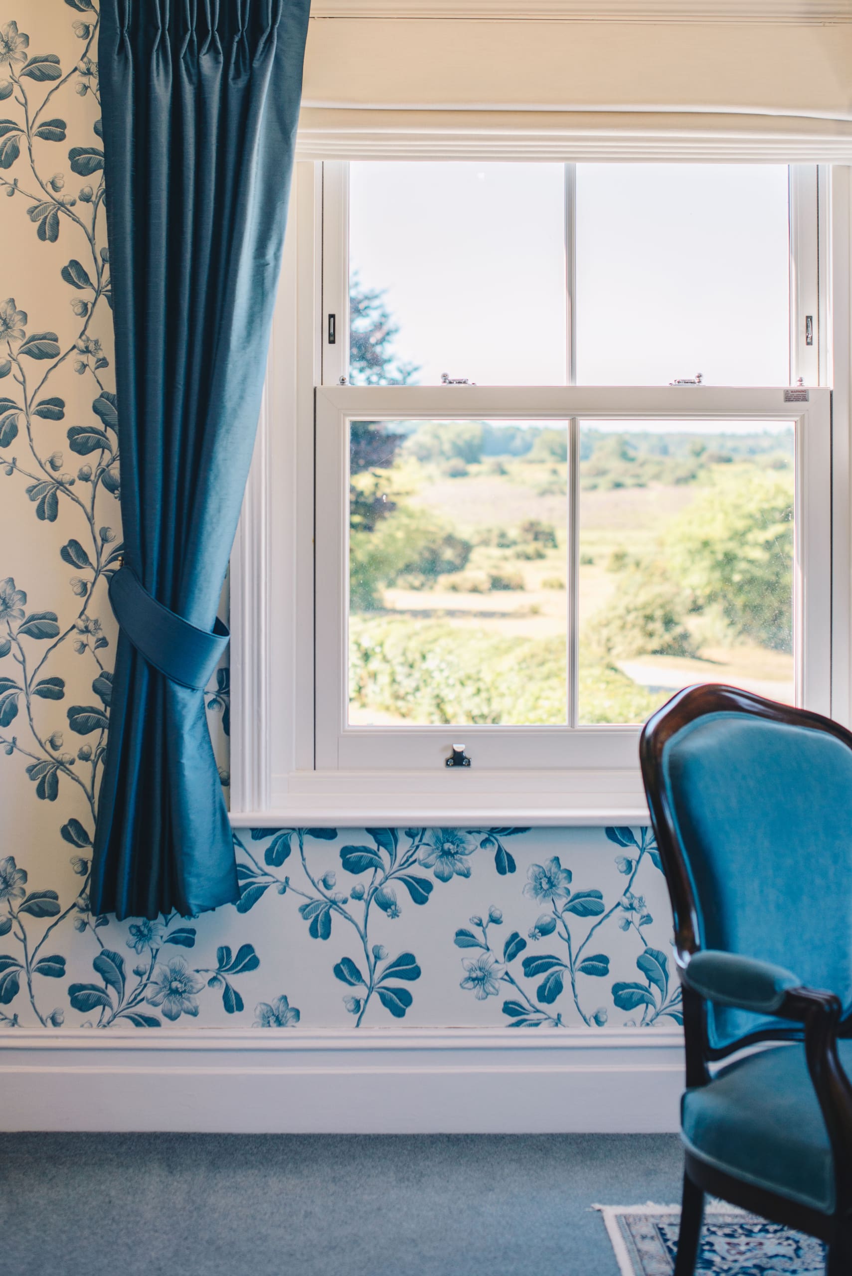 Beautiful traditional home with blue rococo style armchairs and sash windows complete with blue curtains and floral wallpaper.