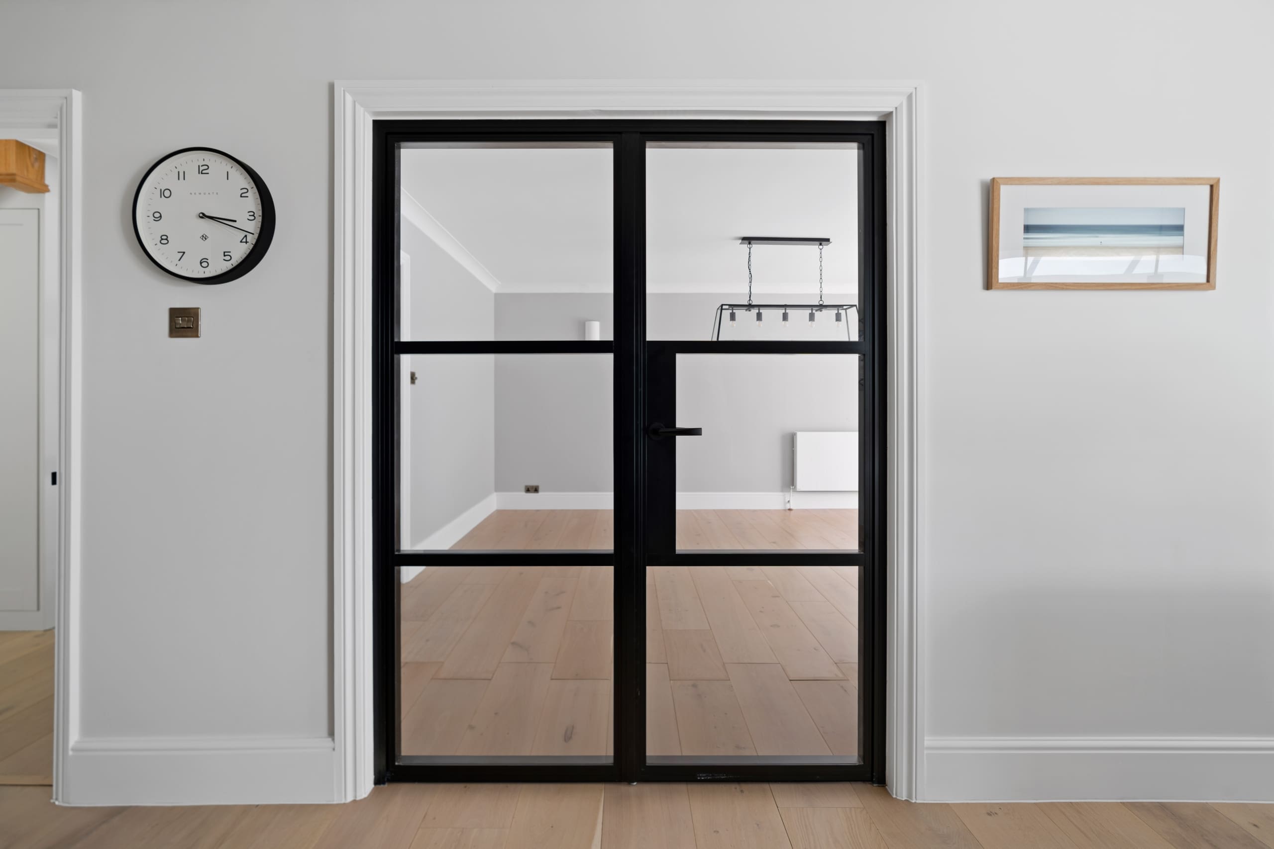 Aluminium black internal french doors with modern clock to the left of the frame hanging on a light grey wall.