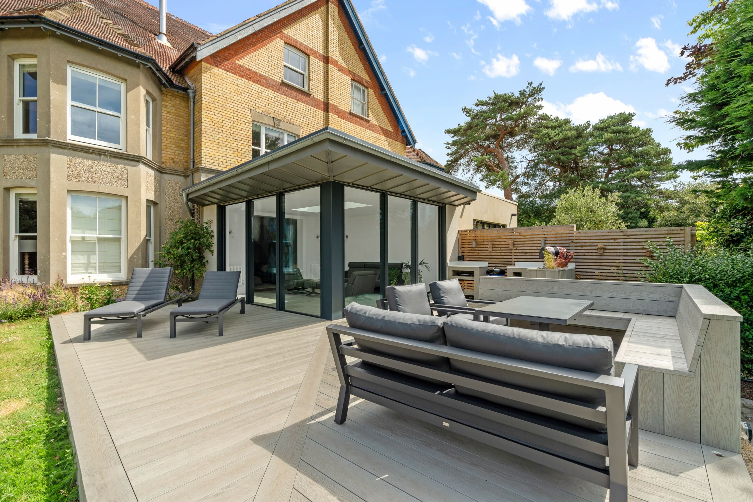 Rear elevation of a substantial home with stunning lightwood decking and garden furniture and modern orangery with sliding doors to each side.
