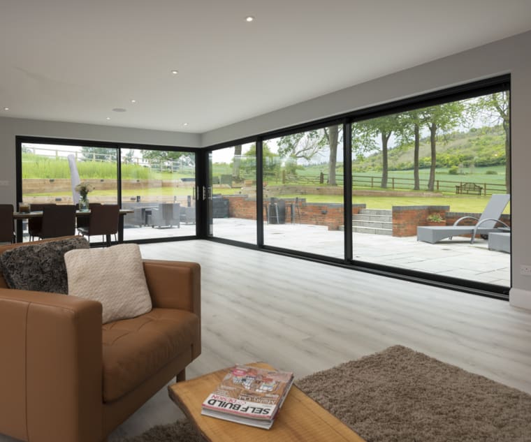 Living room and dining area with dual aspect sliding aluminium doors, with wood flooring and view to secluded gardens overlooking green landscape.